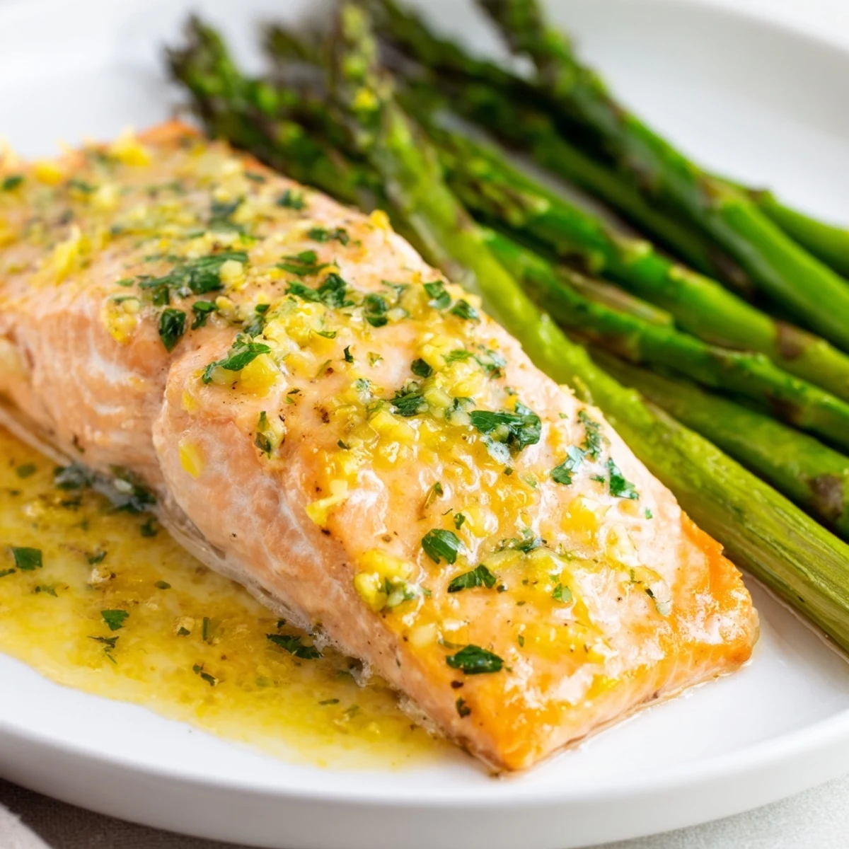 Golden-brown Lemon Garlic Butter Salmon fillets rest beside vibrant green roasted asparagus spears on a white ceramic platter.