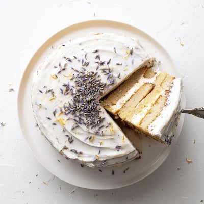 A close-up view of the London Fog Cake with Earl Grey & Lavender reveals textured cake layers and smooth vanilla frosting, with a hint of lemon zest garnish.